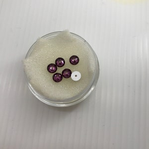 May include: A small container filled with white foam holds six purple round beads and one white round bead.
