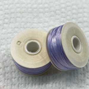 May include: Two spools of lavender-colored thread are positioned on a white surface. The thread is tightly wound around a light beige spool. The spools are close together, with one slightly in front of the other.