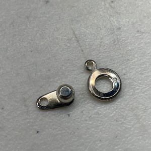 May include: Two silver metal jewelry findings. One is a small, round ring with a loop at the top. The other is a small, flat piece with a loop at one end and a round ball at the other.