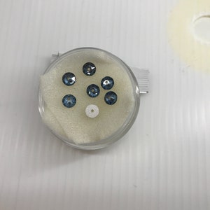 May include: A small clear container with a white foam insert holds seven blue iridescent glass rhinestones.
