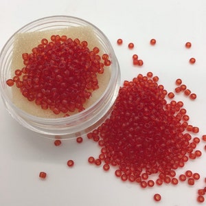 Puede incluir: Un pequeño recipiente transparente lleno de cuentas de semillas rojas. El recipiente está abierto y algunas cuentas se han derramado sobre una superficie blanca.