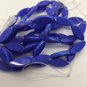 Op de afbeelding: Een streng blauwe glazen kralen met een parelmoer afwerking. De kralen zijn ovaalvormig en geregen op een wit draad.