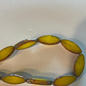 Op de afbeelding: Een streng gele ovale glazen kralen met een bruine en blauwe iriserende afwerking.