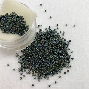 May include: A collection of small, iridescent, dark green and blue seed beads. The beads are scattered on a white surface, with some contained in a clear plastic jar with a white foam insert. These beads are ideal for jewelry making and crafts.