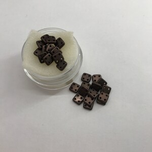 Puede incluir: Un pequeño recipiente lleno de cuentas cuadradas marrones con agujeros. Las cuentas están esparcidas sobre una superficie blanca.