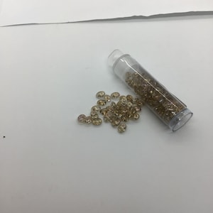 May include: A clear plastic tube filled with small, round, iridescent gold beads. The beads are scattered on a white surface.