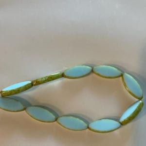 Op de afbeelding: Een streng lichtblauwe ovale kralen met een groene rand. De kralen zijn geregen op een dunne draad.