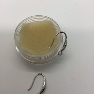Puede incluir: Dos ganchos para pendientes de plata, uno en un pequeño recipiente con una esponja beige y el otro sobre una superficie blanca.