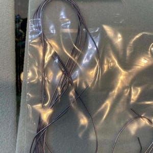 May include: A bundle of thin, metallic silver-toned French wire or gimp cord contained within a clear plastic zip-top bag. The bag features a white adhesive label with the text French Wire/Soft Hevielle and a barcode printed on the surface.