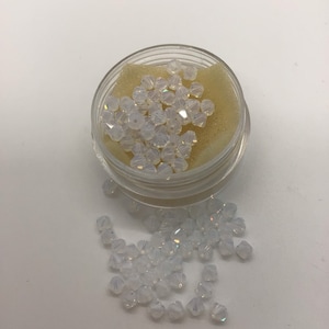 May include: A small container filled with white iridescent glass beads. The beads are scattered on a white surface.