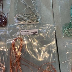 May include: Coils of metallic wire in silver and copper tones, each individually packaged in clear plastic bags. The labels on the bags read French Bull Wire Silver and French Wire Soft Copper, with visible barcodes and pricing information included.