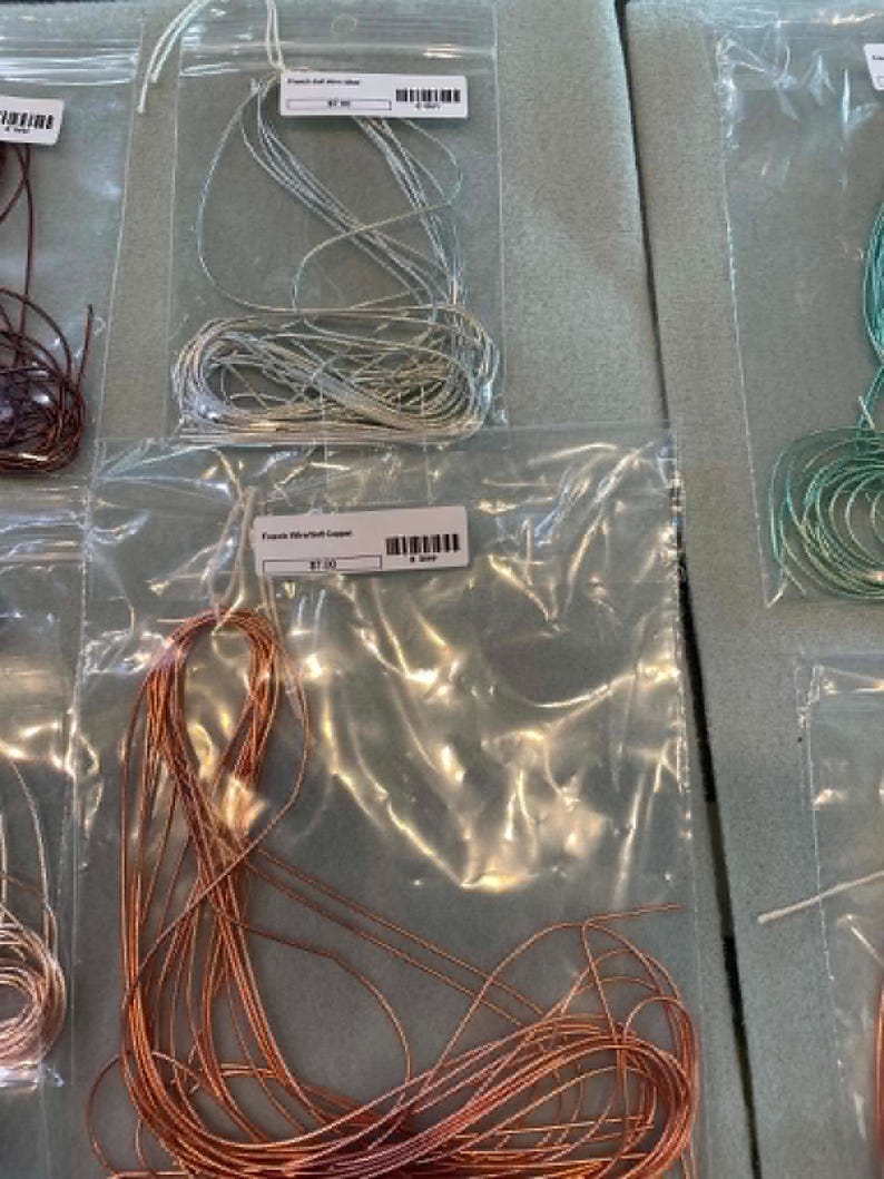 May include: Coils of metallic wire in silver and copper tones, each individually packaged in clear plastic bags. The labels on the bags read French Bull Wire Silver and French Wire Soft Copper, with visible barcodes and pricing information included.