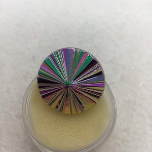 May include: A round, iridescent button with a starburst design in shades of green, purple, and gold. The button is sitting in a clear plastic container with a beige foam insert.