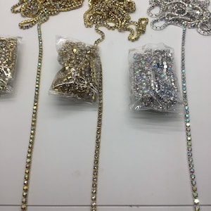 Può includere: Tre rotoli di catena di strass in oro, argento e colori iridescenti. Ogni rotolo è confezionato in un sacchetto di plastica trasparente. Le catene sono disposte su una superficie bianca.