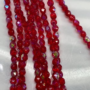 May include: Multiple strands of faceted, deep red glass beads arranged vertically. Each bead features a reflective, iridescent finish that catches the light, creating a shimmering effect across the surface of the small, round, uniform glass components.
