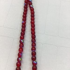 Op de afbeelding: Een ketting of armband van rode, ronde kralen met een iriserend effect. De kralen zijn aan elkaar geregen, waardoor een doorlopende lus ontstaat. De kralen zijn dieprood van kleur en lijken van glas of een vergelijkbaar materiaal te zijn.