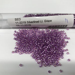May include: A close-up of a small tube of purple glass beads with a label that reads "SED 11-2219 Silverlined Lt. Grape TR-11-2219".