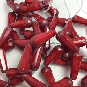 May include: A collection of red glass beads with a teardrop shape and faceted sides. The beads are strung together with clear string, and some have a glittery finish on the rounded end. The beads are scattered on a white surface.