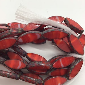 Op de afbeelding: Een streng rode en witte ovale glazen kralen met een gespikkelde afwerking. De kralen zijn geregen op een wit koord.