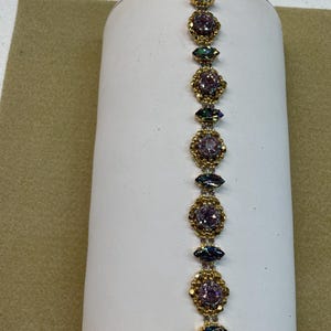Puede incluir: Pulsera dorada con cristales morados e iridiscentes en forma ovalada y de lágrima. La pulsera tiene un cierre decorativo y se exhibe sobre un soporte cilíndrico blanco.