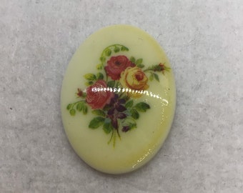 May include: A cream-colored oval-shaped ceramic cabochon with a floral design. The design features three roses in shades of red, yellow, and pink, with green leaves and purple accents.