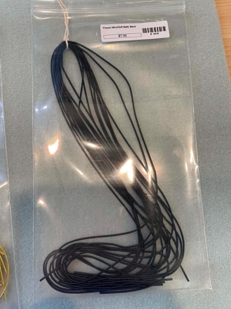 May include: A bundle of thin, black, flexible cord material packaged in a clear plastic bag. The bag features a white adhesive label at the top with the text "French Wire/Gimp Matte Black" and a barcode, along with a small white string loop.