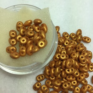 May include: A container of gold-colored seed beads. The beads are oval shaped with two holes in each bead.