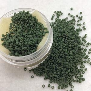 May include: A close-up of a small container filled with dark green seed beads. The beads are scattered on a white surface.