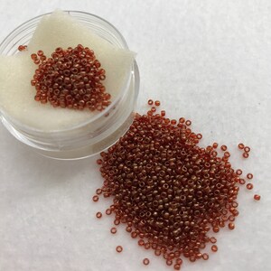 May include: A small clear container filled with a large quantity of small, round, copper-coloured beads. Some beads are scattered on a white surface.