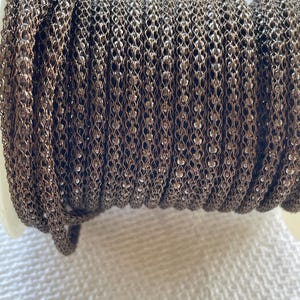 May include: A spool of dark bronze-toned metallic mesh cord featuring a fine, intricate chain-like texture. The cord is tightly wound in multiple parallel rows around a white plastic spool, showcasing a consistent, repeating woven pattern.