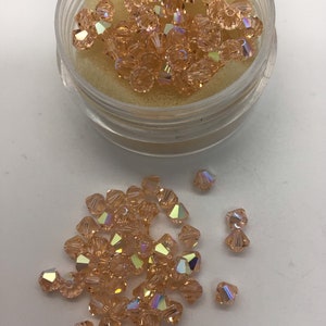 May include: A small container filled with iridescent peach-colored glass beads. The beads are scattered on a white surface.
