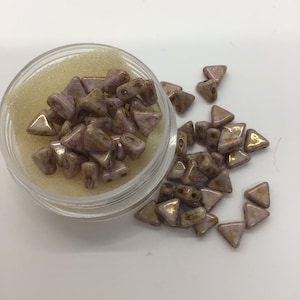 Może przedstawiać: Mały pojemnik wypełniony jasnoróżowymi i złotymi szklanymi koralikami w kształcie trójkątów. Koraliki są rozrzucone wokół pojemnika na białym tle.