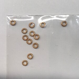 May include: A close-up of a clear plastic bag containing a group of small, gold-colored jump rings. The jump rings are scattered randomly in the bag.