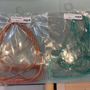 May include: Two clear plastic bags containing thin metallic wire. The left bag holds copper-colored wire, and the right bag holds teal-colored wire. Each bag features a white rectangular label with a barcode and text at the top center.