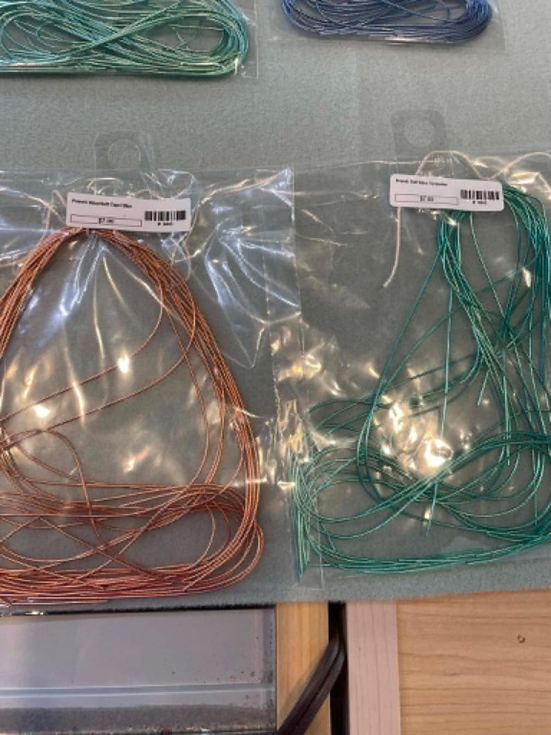 May include: Two clear plastic bags containing thin metallic wire. The left bag holds copper-colored wire, and the right bag holds teal-colored wire. Each bag features a white rectangular label with a barcode and text at the top center.