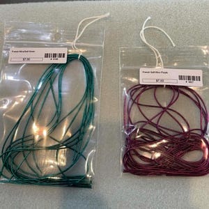 May include: Two clear plastic bags containing thin, metallic-colored cord. The left bag holds dark green cord labeled French Wire/Soft Green. The right bag holds dark purple cord labeled French Soft Wire Purple. Both bags feature white hanging loops.