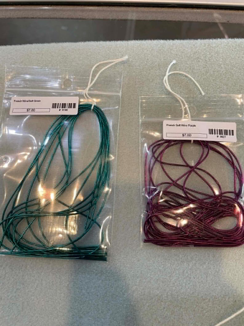 May include: Two clear plastic bags containing thin, metallic-colored cord. The left bag holds dark green cord labeled French Wire/Soft Green. The right bag holds dark purple cord labeled French Soft Wire Purple. Both bags feature white hanging loops.