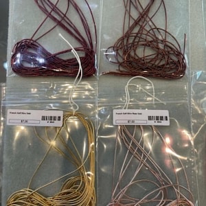 May include: Four clear plastic bags containing coiled French wire in burgundy, brown, gold, and rose gold colors. Each bag features a white label with text and a barcode. The wire is thin, metallic, and coiled in a loose, circular arrangement.