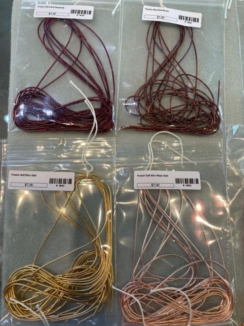 May include: Four clear plastic bags containing coiled French wire in burgundy, brown, gold, and rose gold colors. Each bag features a white label with text and a barcode. The wire is thin, metallic, and coiled in a loose, circular arrangement.