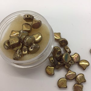 May include: A small container filled with gold-colored glass beads. The beads are shaped like half-moons and have a iridescent finish. Some beads are scattered outside the container.