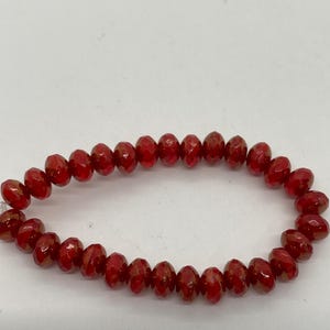 May include: A strand of faceted, oval-shaped beads in a deep red hue. The beads are strung together, forming a circular shape. The beads have a glossy finish and reflect light, creating a sparkling effect. The beads are likely for jewelry making.