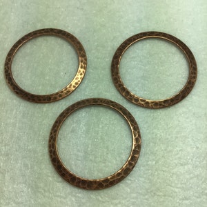 Op de afbeelding: Drie gehamerde koperen ringen, elk met een gladde, ronde opening. De ringen zijn in een driehoekige vorm op een lichtblauw oppervlak gerangschikt.