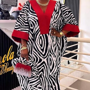 May include: A long, flowing dress with a striking black and white zebra pattern. The dress has a red neckline and sleeve cuffs, paired with a small red beaded clutch. Gold jewellery adds a touch of elegance to the ensemble.
