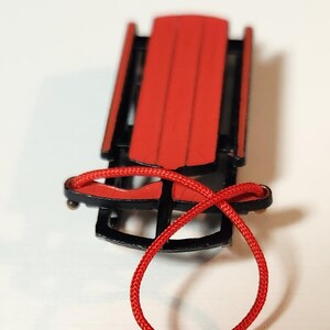 May include: A small red and black sled with a red rope. The sled features a red seat and black runners, a classic design. The red rope is looped. A miniature winter decoration, reminiscent of childhood sledding.