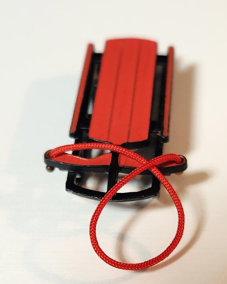May include: A small red and black sled with a red rope. The sled features a red seat and black runners, a classic design. The red rope is looped. A miniature winter decoration, reminiscent of childhood sledding.