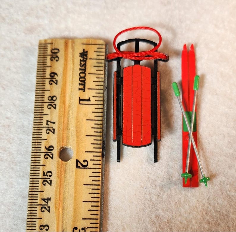 May include: Miniature red sled with black trim and a red rope, next to a pair of red skis with green accents and silver poles. The sled is approximately 2.5 cm long. A wooden ruler is in the image.