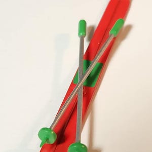 May include: A pair of red toy skis with green and silver ski poles. The skis have a green accent and are angled. The ski poles have green tips and are crossed over the skis. The skis are a toy.
