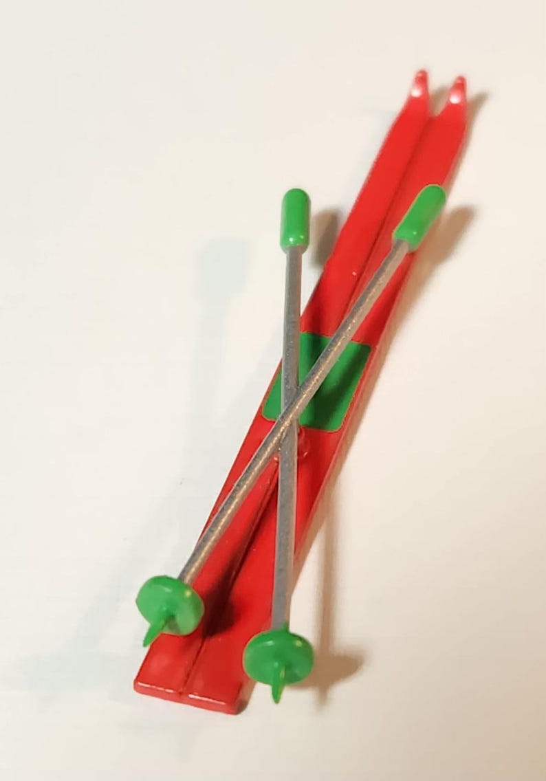 May include: A pair of red toy skis with green and silver ski poles. The skis have a green accent and are angled. The ski poles have green tips and are crossed over the skis. The skis are a toy.