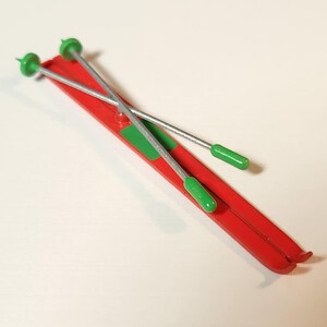 May include: A pair of red toy skis with green accents and silver ski poles. The skis have a green rectangle near the centre. The ski poles have green handles and tips. The toy is likely for a child.