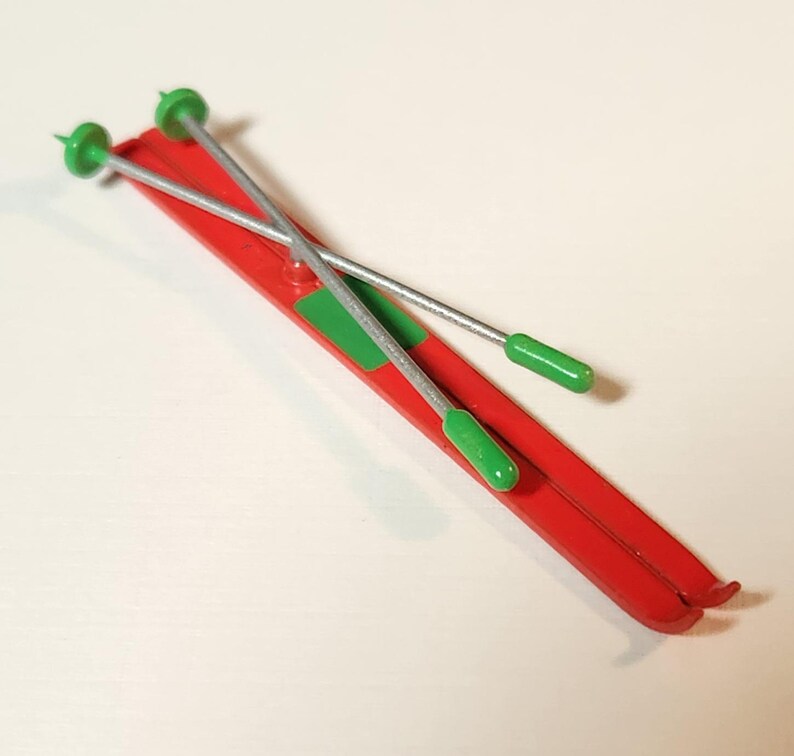 May include: A pair of red toy skis with green accents and silver ski poles. The skis have a green rectangle near the centre. The ski poles have green handles and tips. The toy is likely for a child.